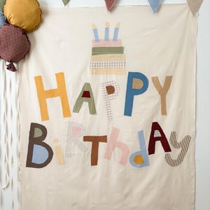 May include: A cream-colored fabric wall hanging featuring the appliqued text HAPPY BIRTHDAY in various patterned fabrics, a three-tiered cake with three candles, and a cluster of fabric balloons with ribbons, topped by a row of triangular bunting.