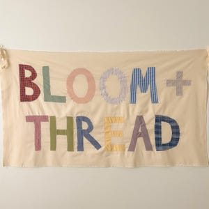 May include: A rectangular off-white fabric banner featuring the text BLOOM + THREAD appliqued in various patterned and solid-colored fabric letters. The letters include red plaid, solid green, pink, light blue stripes, and navy blue plaid.