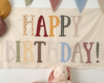 Peut inclure: Une bannière en tissu crème avec le texte HAPPY BIRTHDAY en lettres de tissu à motifs multicolores. Au-dessus de la bannière est suspendue une guirlande de fanions triangulaires de différentes couleurs. Un cochon et un lapin en peluche sont assis.