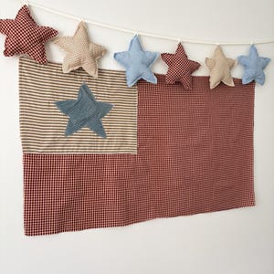 Puede incluir: Decoración de pared patriótica con diseño de bandera. La bandera está hecha de tela a cuadros rojos y blancos y tela a rayas, con una estrella azul. Una guirnalda de estrellas rellenas en varios patrones y colores cuelga arriba.
