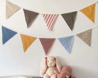 Peut inclure: Une guirlande décorative avec des fanions en tissu colorés est suspendue au-dessus d'une table en bois. Un lapin et un cochon en peluche sont posés sur la table. Les fanions sont de différentes couleurs et motifs, dont le bleu, le jaune et le rouge.