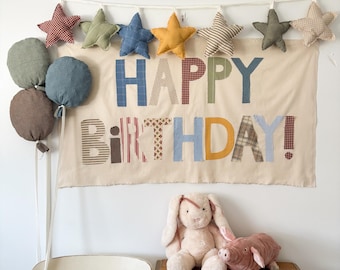 Peut inclure: Une bannière d'anniversaire avec l'inscription "HAPPY BIRTHDAY!" en lettres patchwork colorées. La bannière est ornée d'étoiles et de ballons décoratifs. Un lapin et un cochon en peluche sont posés sur une table en bois.