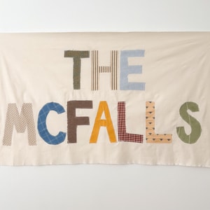 May include: A rectangular off-white fabric banner featuring the text THE MCFALLS appliqued in various patterned and colored fabric letters. The letters include green, brown, blue, yellow, and red gingham, striped, and solid fabric patches.