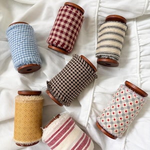 May include: A collection of seven decorative spools with wooden ends and fabric wraps. The fabrics feature various patterns, including gingham, stripes, and stars, in colors like blue, red, and yellow. The spools are arranged on a white, ruffled surface.
