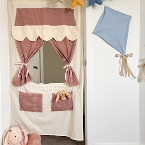May include: A cream-colored playhouse door cover with red gingham accents, scalloped trim, and pockets. A star-shaped garland hangs above, and a blue kite is attached to the wall. Plush toys, including a bunny and pig, sit on the floor.