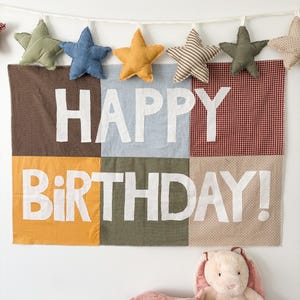 May include: A colorful birthday banner with the words "HAPPY BIRTHDAY!" in white block letters. Above the banner are star-shaped ornaments in various colors. A stuffed pig and bunny are in the foreground.