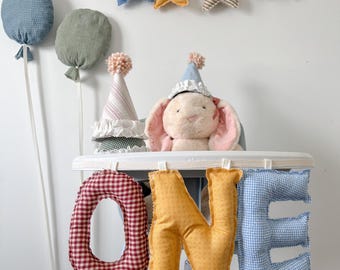 Peut inclure: Une bannière en tissu épelant le mot ONE en grandes lettres rembourrées aux motifs vichy rouge, jaune uni et vichy bleu. Un lapin en peluche portant un chapeau de fête est placé derrière la bannière, avec des décorations en forme d'étoiles et de ballons.