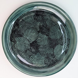 Può includere: Un piatto rotondo in ceramica color verde acqua con finitura lucida. L'interno del piatto presenta un motivo ripetuto di foglie di felce verde scuro stilizzate su sfondo nero. Il bordo del piatto è di una tonalità verde acqua più scura.