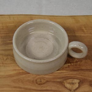 May include: A speckled, off-white ceramic bowl with a rounded shape and a small, circular handle. The bowl sits on a wooden surface, showcasing its handmade quality. The bowl is ideal for serving dips, sauces, or small portions.