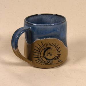 Può includere: Una tazza in ceramica fatta a mano con interno e manico blu e esterno beige. La tazza presenta un'illustrazione nera di una luna crescente, stelle e raggi di sole. La tazza è alta circa 10 cm.