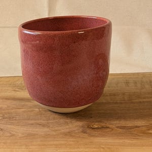 Op de afbeelding: Een handgemaakte keramische mok met een gespikkelde rode glazuur. De mok heeft een afgeronde vorm en een crèmekleurige basis. Hij staat op een houten oppervlak, met een neutrale achtergrond.