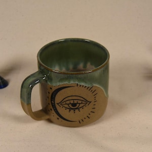 May include: A ceramic mug with a green interior and handle, and a tan exterior featuring a crescent moon and eye design. The mug is accompanied by crystals and a bundle of dried lavender.