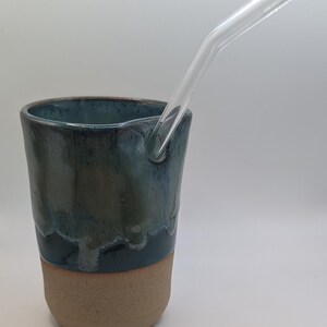 May include: A handcrafted ceramic tumbler with a clear glass straw. The tumbler features a two-tone design, with a teal and green glazed top and a natural tan base. The straw is angled and sits inside the tumbler.