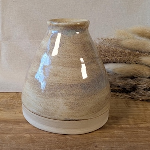 Op de afbeelding: Een handgemaakte keramische vaas met een brede basis en een smalle hals. De vaas heeft een beige en bruin kleurenschema met een glanzende afwerking. De vaas staat op een houten oppervlak.