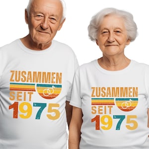 May include: Two people wearing matching white t-shirts. The shirts feature the German text "ZUSAMMEN SEIT 1975" in a retro design with rings. The design includes orange, yellow, and blue stripes.