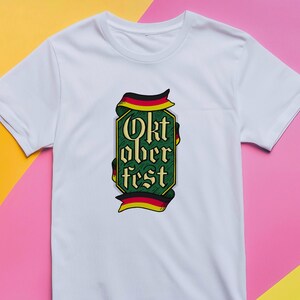 May include: White t-shirt featuring a green and gold Oktoberfest design. The design includes the text "Oktoberfest" in a decorative font, framed by a ribbon in the colors of the German flag.