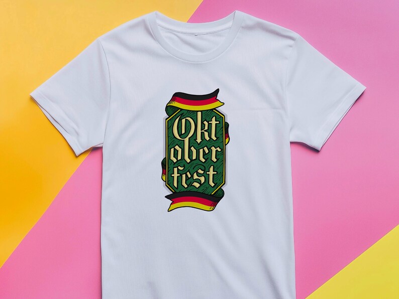 May include: White t-shirt featuring a green and gold Oktoberfest design. The design includes the text "Oktoberfest" in a decorative font, framed by a ribbon in the colors of the German flag.