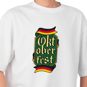 May include: White t-shirt featuring a green and gold Oktoberfest design with the text "Oktoberfest" in a stylized font. The design includes ribbons in the colors of the German flag. A casual, comfortable tee.