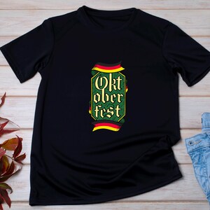 May include: Black t-shirt with a green and yellow Oktoberfest design. The design includes the words "Oktoberfest" in a decorative font, with German flag accents. The shirt is displayed on a light wood surface.
