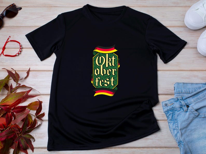 May include: Black t-shirt with a green and yellow Oktoberfest design. The design includes the words "Oktoberfest" in a decorative font, with German flag accents. The shirt is displayed on a light wood surface.