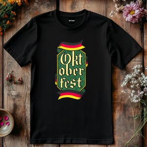 May include: Black t-shirt with the words "Oktoberfest" in a decorative font, framed by a green border and German flag-inspired ribbons. The shirt is made of a soft material and is suitable for casual wear.