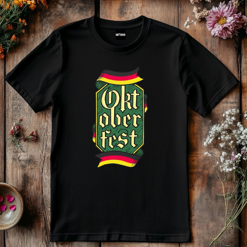 May include: Black t-shirt with the words "Oktoberfest" in a decorative font, framed by a green border and German flag-inspired ribbons. The shirt is made of a soft material and is suitable for casual wear.