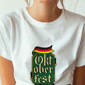May include: White t-shirt featuring a green and gold Oktoberfest design. The design includes the words "Oktoberfest" in a stylized font, with a ribbon in the colors of the German flag.
