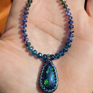 Op de afbeelding: Een ketting met een druppelvormige donkere opaal hanger met levendige groene en blauwe flitsen, omlijst door een kralenrand. De streng bestaat uit gefacetteerde ronde kralen die in kleur verlopen van diepblauw naar heldergroen.