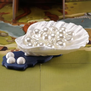 Peut inclure: Un récipient blanc en forme de coquillage rempli de nombreuses perles blanches. La coquille repose sur une base bleu foncé de forme irrégulière. L'arrière-plan présente un plateau de jeu coloré avec un motif de carte.