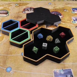 Könnte beinhalten: Schwarze und goldene sechseckige Würfelbehälter mit farbigen Fächern und einem schwarzen Deckel. Mehrere vielseitige Würfel in verschiedenen Farben befinden sich im Inneren des Behälters, der auf einer Oberfläche mit Kartenmotiv platziert ist. Die Behälter sind für Tischspiele konzipiert.