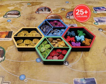 Puede incluir: Un conjunto de tres organizadores hexagonales para juegos de mesa entrelazados con interiores negros y bordes de colores amarillo, azul y verde. Las bandejas contienen varias fichas de juego, incluyendo monedas doradas, discos azules y morados, fichas de hojas verdes, iconos de carne rojos y figuras de dragones.