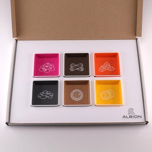 May include: A set of six colorful square game component trays in a cardboard box. Each tray is a different color and contains a white icon representing game resources like berries, logs, and coins. The Albion Craftworks logo is in the bottom right corner.