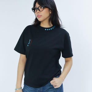 Może przedstawiać: Czarny t-shirt z okrągłym dekoltem, z serią małych, jasnoniebieskich, okrągłych ozdób wokół dekoltu. Koszulka ma również mały wzór na lewej piersi w kolorze czerwonym i jasnoniebieskim. Modelka ma na sobie niebieskie dżinsy i czarne okulary.