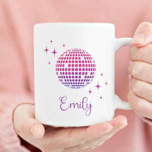 May include: White ceramic mug with a pink and purple disco ball design and the name "Emily" in purple script. The mug has a white handle.