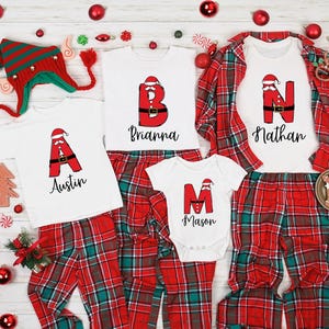 May include: A collection of Christmas pajamas with a red and green plaid pattern. The shirts feature letters designed as Santa Claus, with names below. A matching elf hat and Christmas ornaments complete the festive scene.
