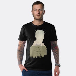 Könnte beinhalten: Schwarzes T-Shirt mit einem Silhouettendesign und dem Text "MAN IS THE CRUELEST ANIMAL". Das Design beinhaltet eine Landschaft und Vögel. Das T-Shirt hat einen Rundhalsausschnitt.