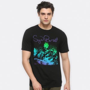 以下が含まれることがあります： ギターを弾いているシド・バレットのカラフルな画像がプリントされた黒いTシャツ。画像は緑、青、紫のグラデーションで表現されています。画像の上に紫色の文字で「Syd Barrett」と書いてあります。