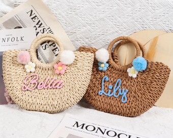 May include: Two straw tote bags with pom poms and embroidered names, "Bella" and "Lily". The bags are light brown and beige with pink and blue flowers.