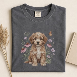 Puede incluir: Camiseta gris oscuro con una ilustración en acuarela de un cachorro marrón y blanco sentado entre flores rosas y moradas y una mariposa. La camiseta tiene una etiqueta Comfort Colors.