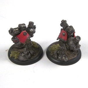 May include: Two miniature metal robots with red accents and a steampunk aesthetic. The robots are standing on black bases with green and brown textured surfaces.