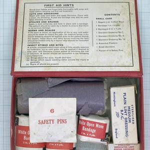 May include: A vintage red first aid kit, open to reveal bandages, cotton wool, and dressings. The kit includes a packet of safety pins and instructions for treating injuries. The text "SAFETY PINS" is visible on a packet.