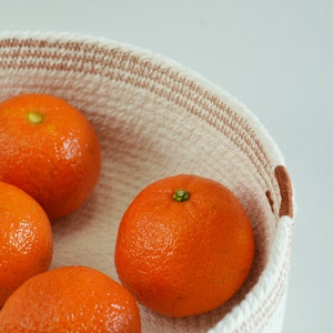 May include: Four whole oranges in a white and brown woven basket. The oranges are arranged in a circle with the tops of the oranges facing the viewer.