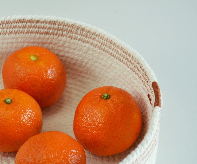 May include: Four whole oranges in a white and brown woven basket. The oranges are arranged in a circle with the tops of the oranges facing the viewer.