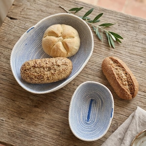 Op de afbeelding: Twee ovale manden gemaakt van opgerold touw met een lichtblauw en wit patroon. De grotere mand bevat een rond broodje en een langwerpig broodje met zaden, terwijl de kleinere mand leeg is. Een takje met groene bladeren ligt in de buurt.