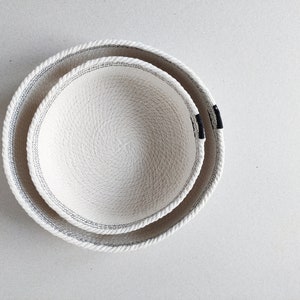 May include: Two white rope bowls, one nested inside the other. The bowls have a black trim around the edge.