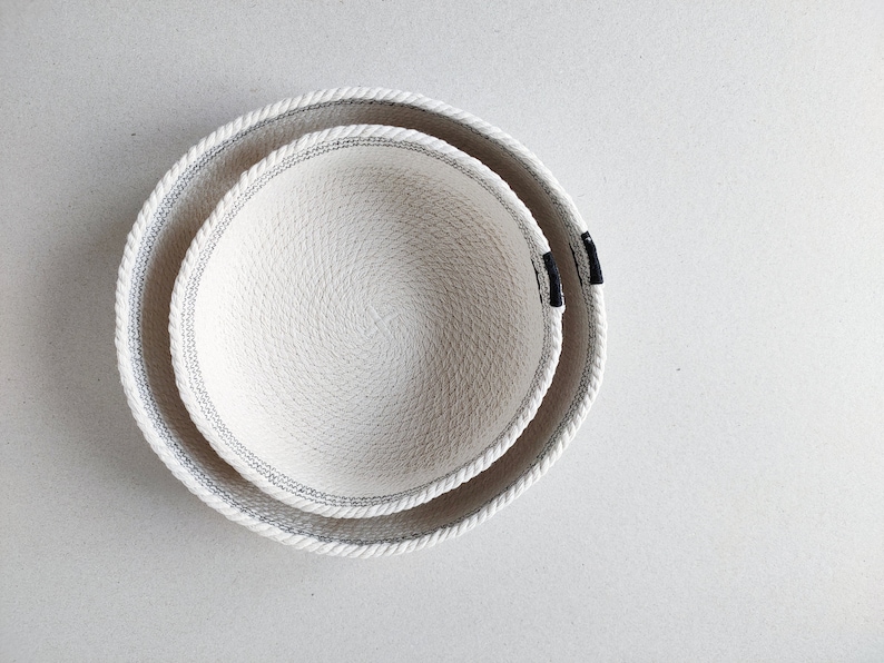 May include: Two white rope bowls, one nested inside the other. The bowls have a black trim around the edge.