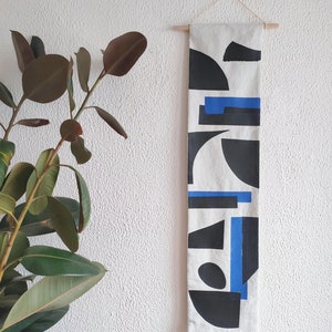 Può includere: Un'appendice da parete in tela bianca con un design geometrico con forme nere e blu. L'appendice è fissata a un'asta di legno.