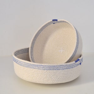 May include: Two white rope baskets with blue trim. The smaller basket is inside the larger basket.