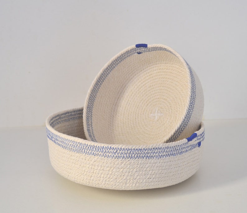 May include: Two white rope baskets with blue trim. The smaller basket is inside the larger basket.