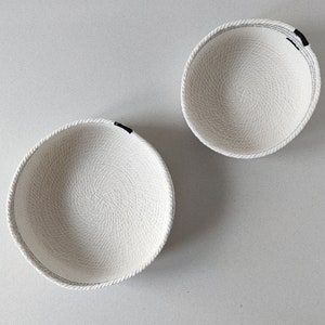May include: Two white rope bowls with black trim. The bowls are empty and sitting on a white surface.